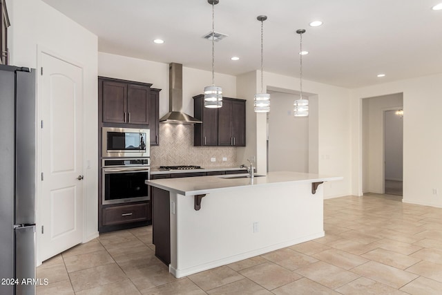 kitchen with appliances with stainless steel finishes, decorative light fixtures, a breakfast bar area, a kitchen island with sink, and wall chimney range hood