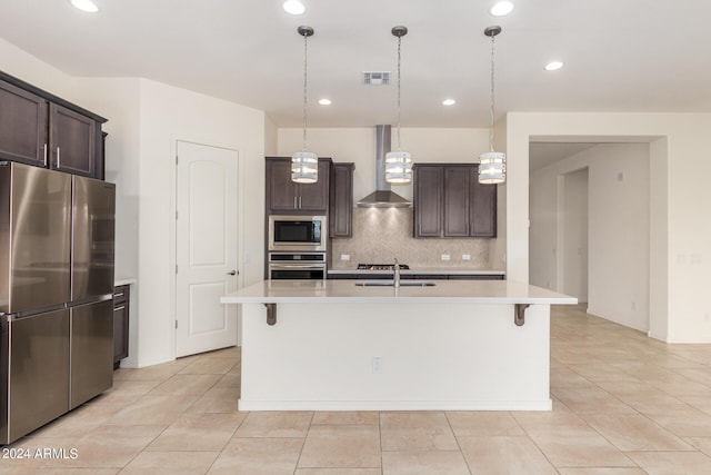 kitchen with wall chimney range hood, stainless steel appliances, hanging light fixtures, and a center island with sink