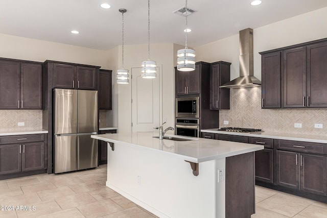 kitchen featuring sink, appliances with stainless steel finishes, an island with sink, pendant lighting, and wall chimney range hood