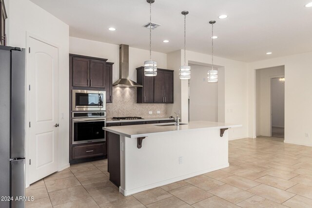 kitchen featuring a breakfast bar, pendant lighting, wall chimney range hood, stainless steel appliances, and a center island with sink