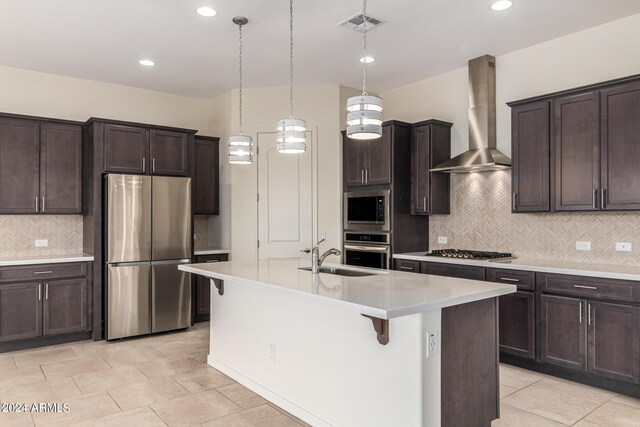 kitchen with appliances with stainless steel finishes, sink, hanging light fixtures, a kitchen island with sink, and wall chimney range hood