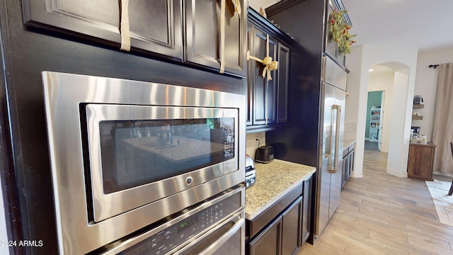 kitchen with built in appliances, dark brown cabinetry, light stone countertops, and light hardwood / wood-style flooring