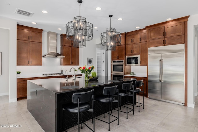 kitchen featuring pendant lighting, wall chimney range hood, built in appliances, a notable chandelier, and a center island with sink