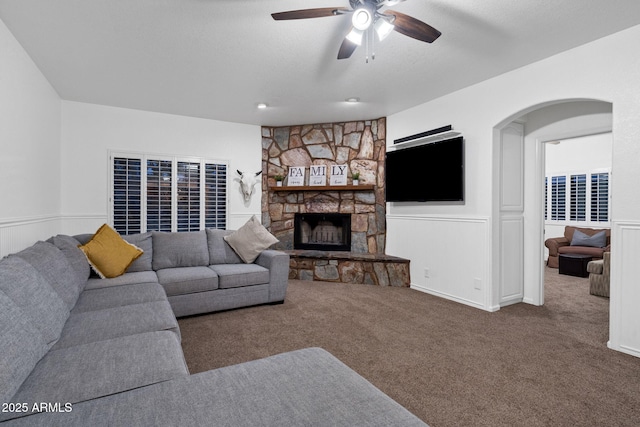carpeted living room with a fireplace, a ceiling fan, a wainscoted wall, and arched walkways