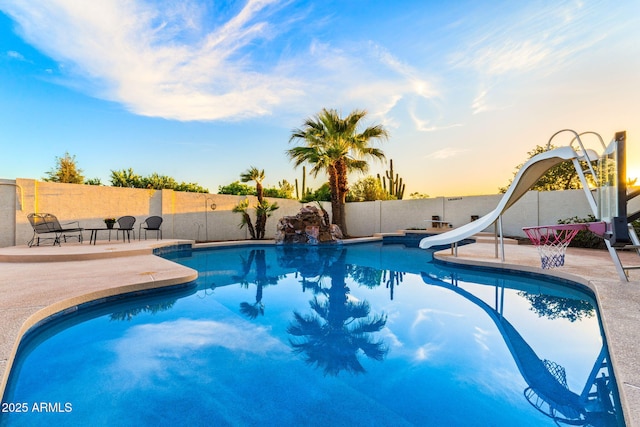 pool at dusk featuring a water slide, a patio, a fenced backyard, and a fenced in pool