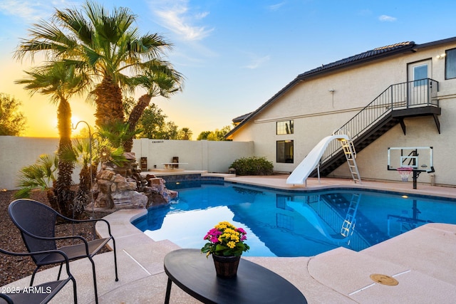 pool at dusk featuring a patio, a fenced in pool, a fenced backyard, stairs, and a water slide