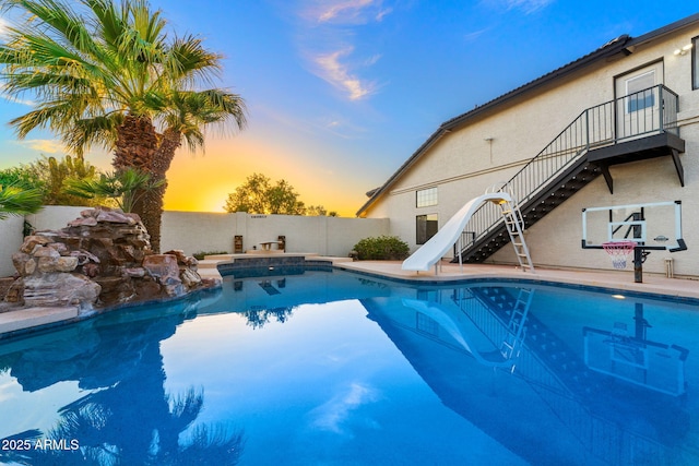 pool at dusk featuring a water slide, a fenced backyard, a fenced in pool, and stairs
