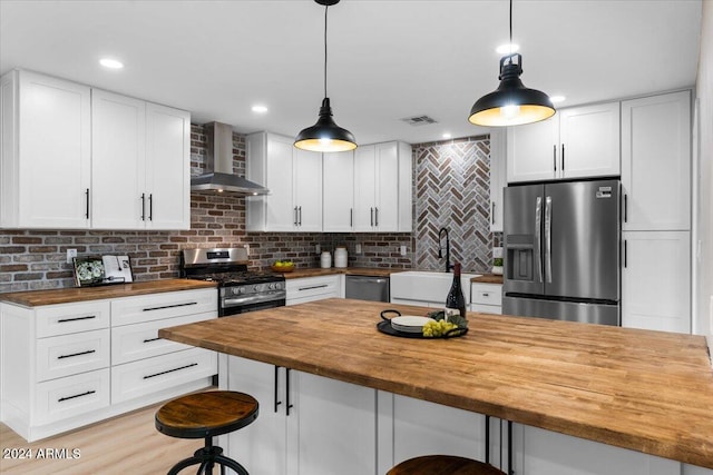 kitchen with stainless steel appliances, wooden counters, wall chimney range hood, white cabinetry, and pendant lighting