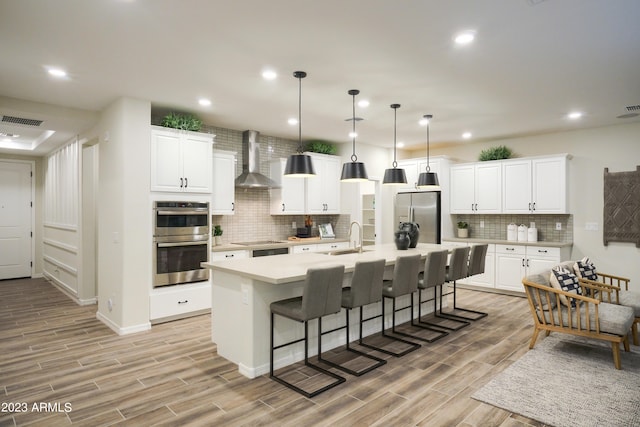 kitchen with appliances with stainless steel finishes, wall chimney exhaust hood, a center island with sink, and light hardwood / wood-style flooring