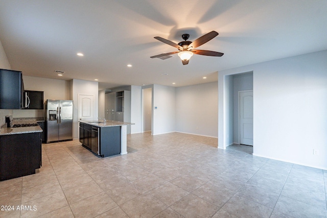 kitchen with light stone counters, ceiling fan, sink, appliances with stainless steel finishes, and a center island with sink