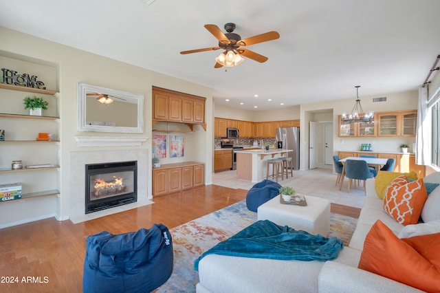 living room featuring a tiled fireplace, built in features, ceiling fan with notable chandelier, and light wood-type flooring