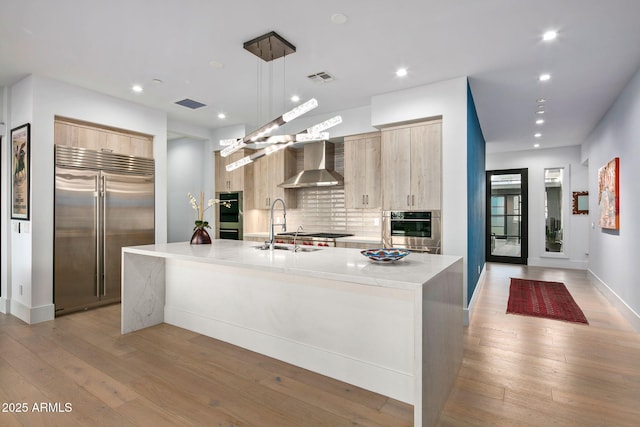 kitchen with light brown cabinetry, hanging light fixtures, stainless steel appliances, a center island with sink, and wall chimney exhaust hood