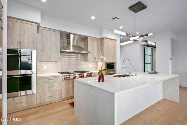 kitchen with appliances with stainless steel finishes, sink, light brown cabinets, a kitchen island with sink, and wall chimney range hood