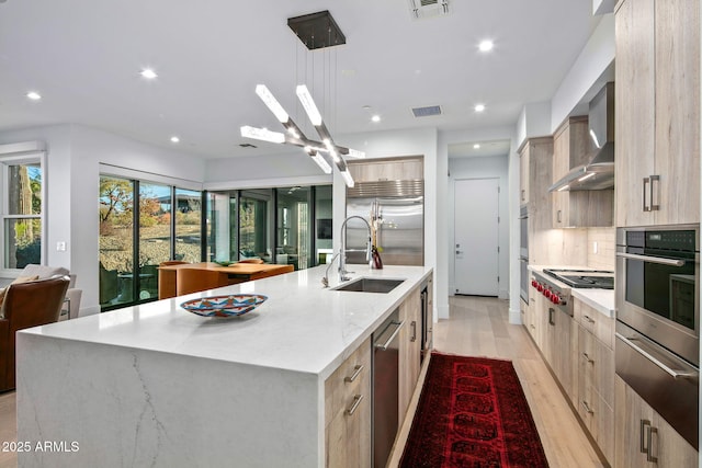 kitchen with wall chimney range hood, sink, stainless steel appliances, a spacious island, and decorative light fixtures