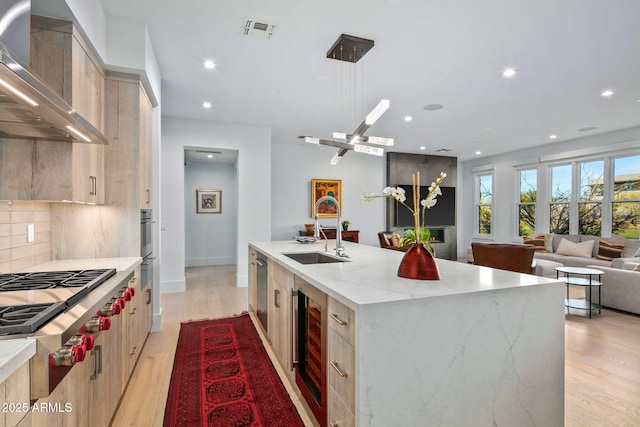 kitchen with wall chimney exhaust hood, light brown cabinetry, sink, an island with sink, and beverage cooler