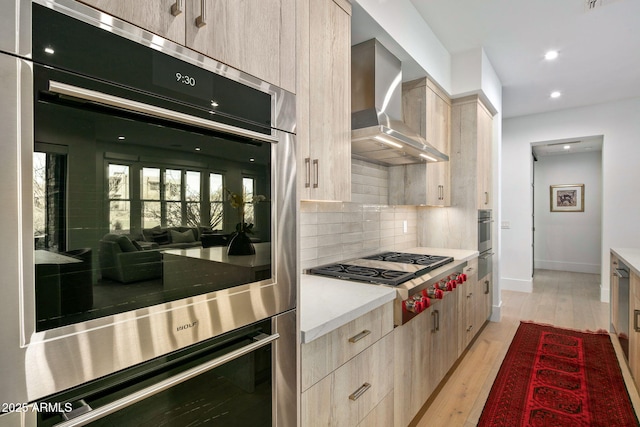 kitchen featuring stainless steel appliances, light brown cabinets, decorative backsplash, wall chimney exhaust hood, and light wood-type flooring