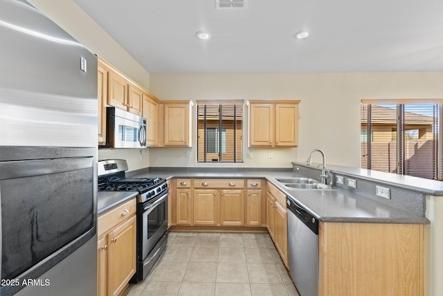 kitchen featuring dark countertops, appliances with stainless steel finishes, a peninsula, light brown cabinetry, and a sink