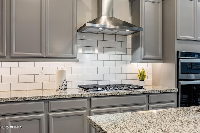 kitchen featuring light stone counters, stainless steel appliances, backsplash, gray cabinetry, and wall chimney exhaust hood