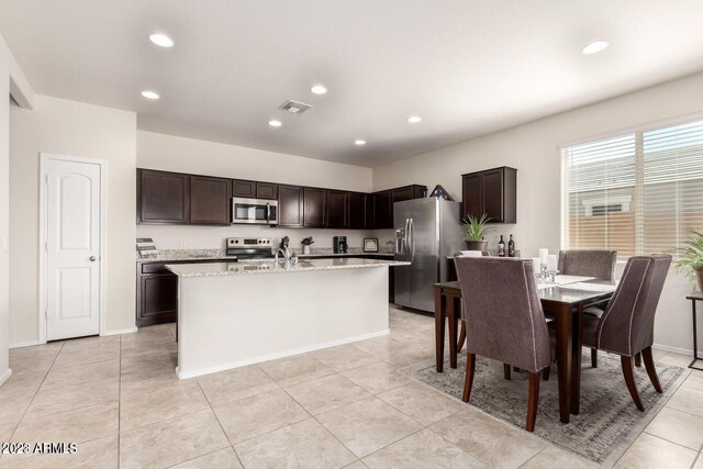 kitchen with light tile patterned floors, stainless steel appliances, light stone countertops, sink, and a center island with sink