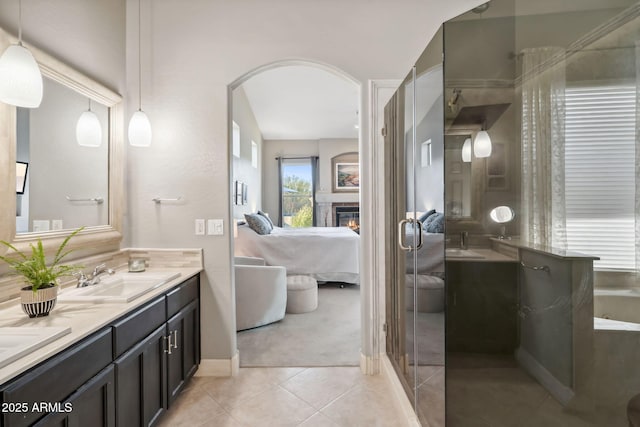 ensuite bathroom featuring a glass covered fireplace, tile patterned flooring, double vanity, a tub, and a stall shower