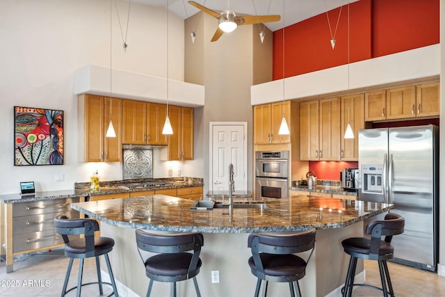 kitchen with stainless steel appliances, ceiling fan, a high ceiling, brown cabinetry, and an island with sink