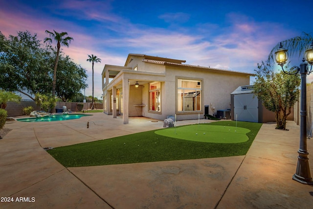 back house at dusk with a storage shed, a patio area, central air condition unit, ceiling fan, and a fenced in pool