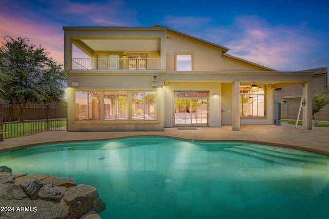 back house at dusk featuring a fenced in pool, a balcony, ceiling fan, and a patio area