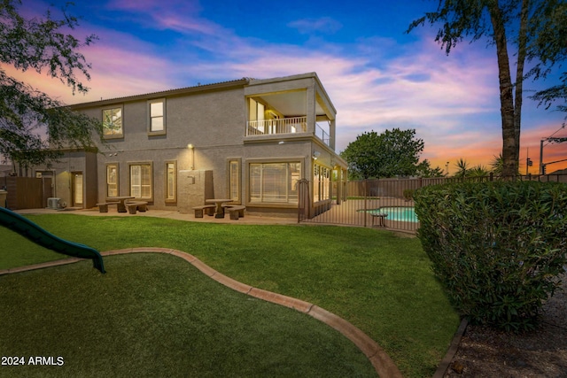 back house at dusk with a balcony, a fenced in pool, a patio area, and a lawn