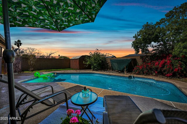 pool at dusk with a storage shed, a fenced in pool, a patio, a fenced backyard, and an outbuilding