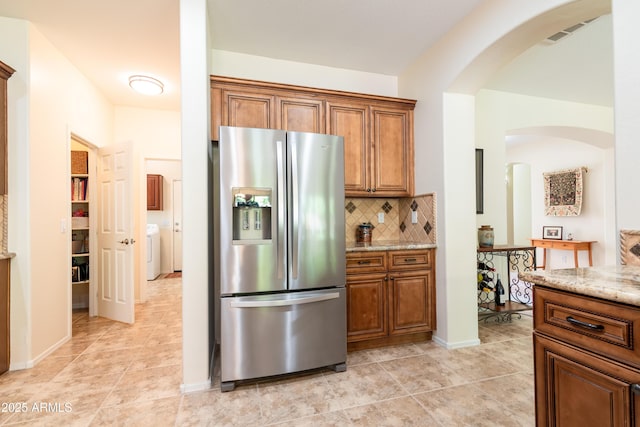 kitchen with brown cabinets, stainless steel refrigerator with ice dispenser, light tile patterned floors, decorative backsplash, and light stone countertops