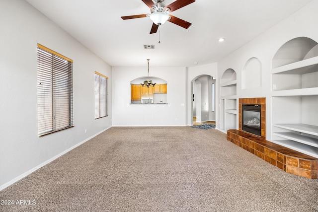 unfurnished living room featuring carpet, built in shelves, ceiling fan, and a tiled fireplace