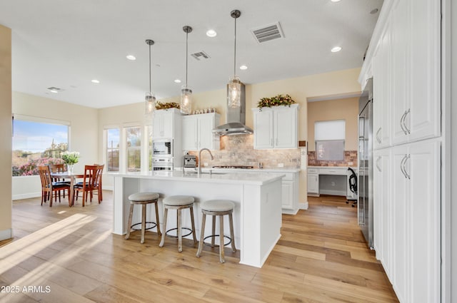 kitchen with a center island with sink, white cabinetry, hanging light fixtures, and wall chimney range hood