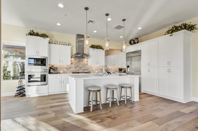 kitchen featuring built in appliances, a center island with sink, white cabinetry, and wall chimney range hood