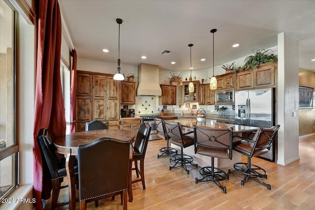 kitchen with custom exhaust hood, a kitchen island with sink, light wood-type flooring, light stone countertops, and appliances with stainless steel finishes