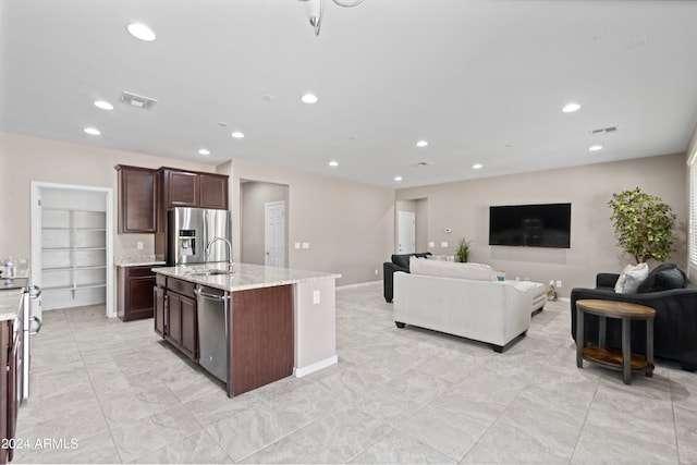 kitchen with sink, dark brown cabinets, a center island with sink, stainless steel appliances, and light stone countertops