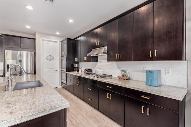 kitchen featuring sink, appliances with stainless steel finishes, dark brown cabinets, light stone countertops, and light wood-type flooring