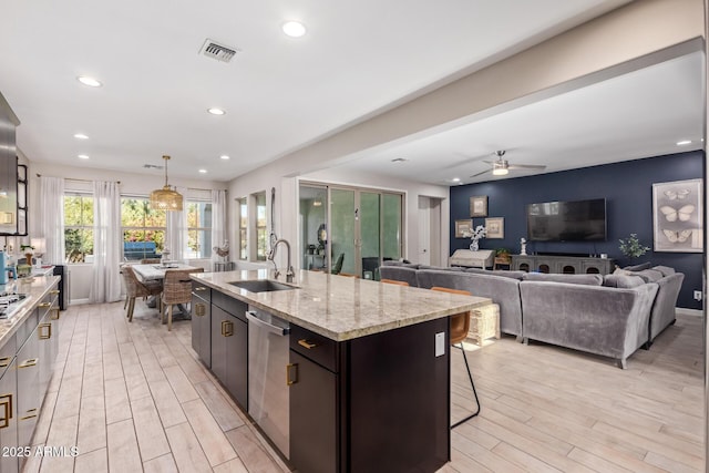 kitchen with sink, hanging light fixtures, a kitchen island with sink, stainless steel dishwasher, and light stone counters