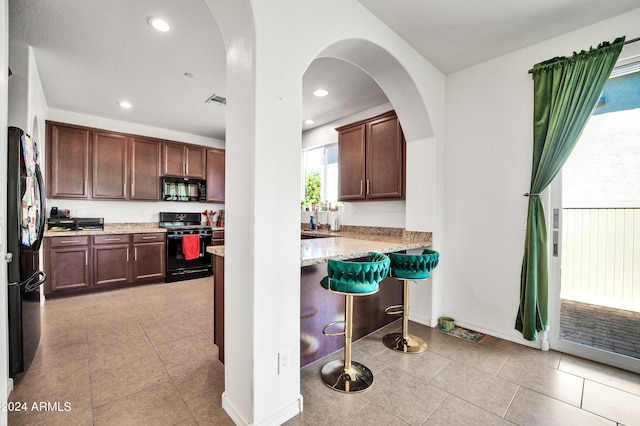 kitchen featuring light tile patterned floors, light stone counters, black appliances, kitchen peninsula, and a breakfast bar area