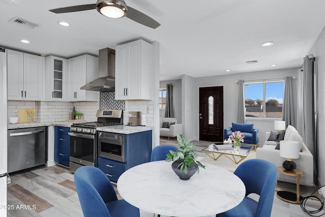 kitchen featuring white cabinetry, light stone counters, stainless steel appliances, blue cabinetry, and wall chimney range hood