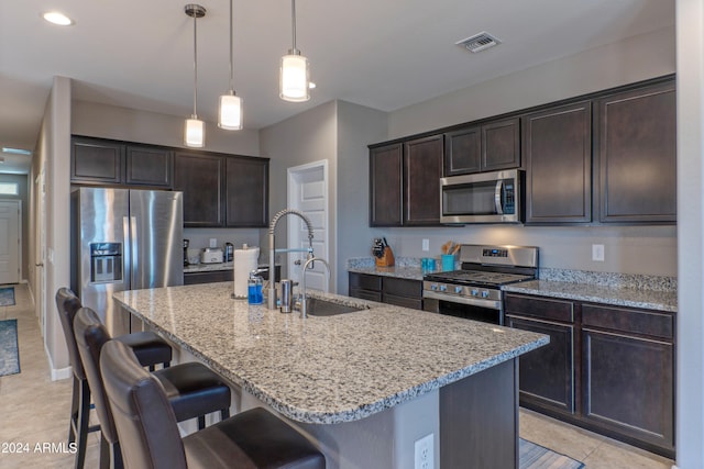 kitchen with pendant lighting, appliances with stainless steel finishes, a kitchen island with sink, light tile patterned floors, and dark brown cabinetry