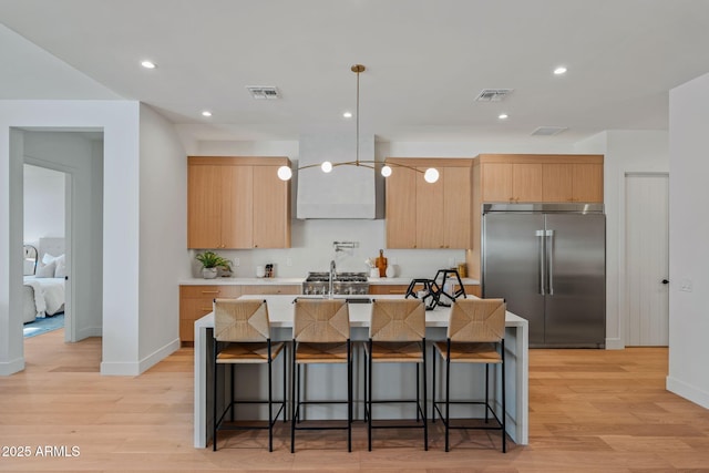 kitchen featuring built in refrigerator, visible vents, a center island with sink, and light brown cabinets