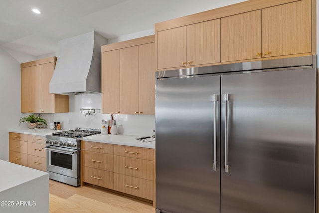 kitchen with light countertops, high end appliances, light brown cabinets, and wall chimney range hood