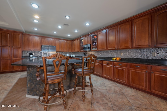 kitchen with a breakfast bar area, dark stone countertops, light tile patterned floors, appliances with stainless steel finishes, and an island with sink