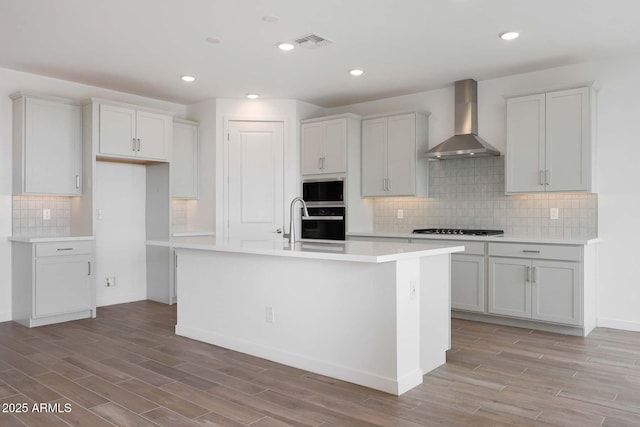 kitchen featuring wall chimney exhaust hood, light wood-type flooring, gas cooktop, stainless steel microwave, and a kitchen island with sink