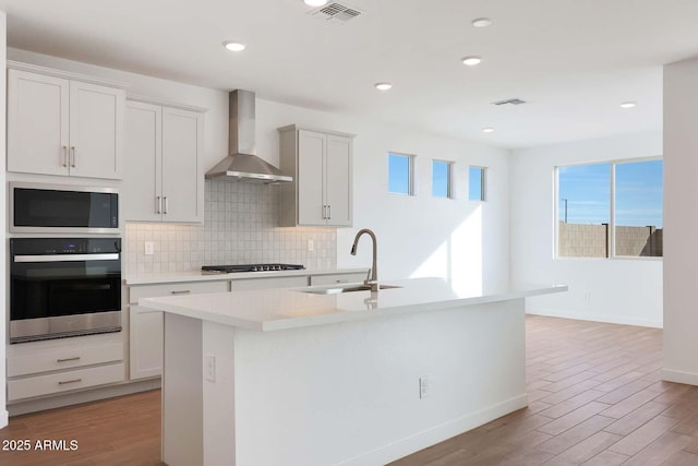 kitchen with sink, appliances with stainless steel finishes, a kitchen island with sink, white cabinetry, and wall chimney exhaust hood