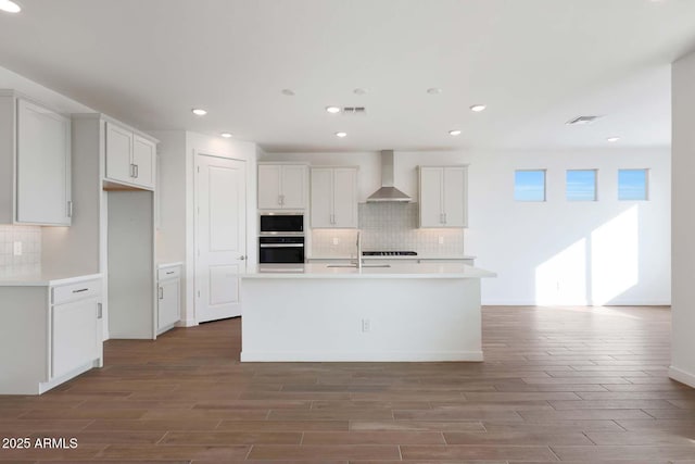 kitchen featuring built in microwave, wall chimney exhaust hood, white cabinetry, an island with sink, and oven