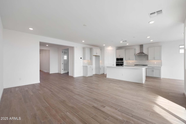 kitchen with backsplash, an island with sink, wall chimney exhaust hood, and light wood-type flooring