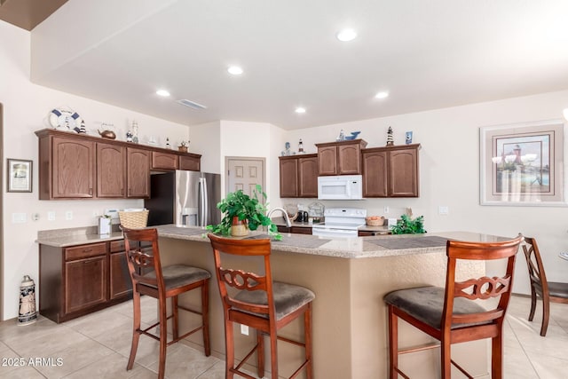 kitchen with light tile patterned flooring, a kitchen breakfast bar, dark brown cabinetry, a center island with sink, and white appliances