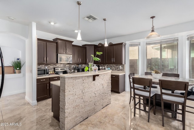 kitchen featuring pendant lighting, tasteful backsplash, a kitchen island with sink, stainless steel appliances, and dark brown cabinets