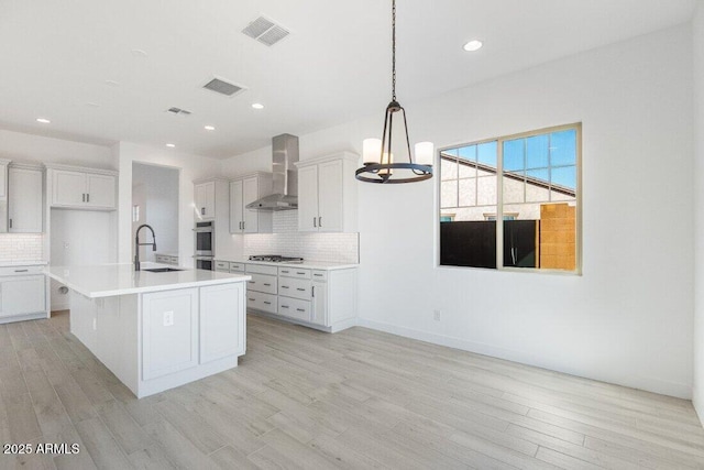 kitchen with white cabinets, a kitchen island with sink, hanging light fixtures, wall chimney exhaust hood, and sink